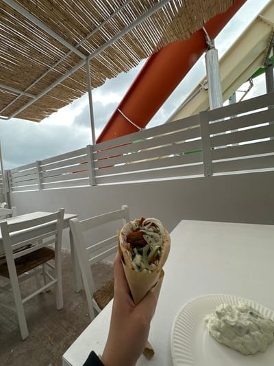 The image shows a low-angle view, looking up at an outdoor eating area, likely in Faliraki, Greece, given the subject matter. A hand holds a gyros wrap, which is the main subject, filled with meat, vegetables, and sauce, partially unwrapped in brown paper. To the right, there is a white paper plate with a dollop of tzatziki sauce.
The environment consists of outdoor structures, including a bamboo-covered section, a reddish-orange pipe, and a white pergola-like structure with evenly spaced slats. Beneath the pergola, white chairs and tables are visible. The sky peeks through the structures, suggesting an overcast day. The flooring under the tables appears textured and worn. No people are visible besides the hand holding the gyros. The scene suggests a casual dining setting.