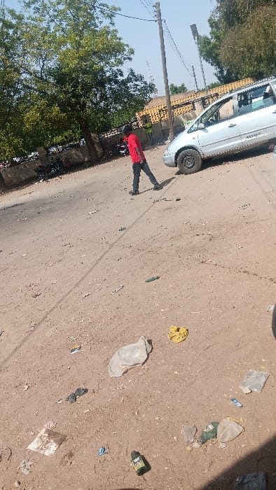 The image captures an outdoor scene on an unpaved, reddish-brown dirt road in Langtang, Nigeria, under clear daylight. A single male individual, wearing a red short-sleeved shirt, dark trousers, and dark footwear, is observed walking from left to right in the midground. To the right of the individual, a light-colored minivan, possibly silver, is parked or stationary, facing left on the dirt surface; the front wheel visible on the left lacks a hubcap and features a black steel rim. The ground surface in the foreground and midground is extensively littered with various items, including multiple plastic bags (one large grey, one smaller clear), a yellow piece of fabric or plastic, scattered paper debris, a dark green bottle with a yellow label, and other small, indistinguishable objects. In the background, green-leafed trees are visible on the left, while a low, light-brown building with horizontal textured patterns stands behind the vehicle. A wooden utility pole with overhead lines extends vertically near the center-right, and a lattice communication tower is faintly visible further in the distance. The sky is clear and light blue.