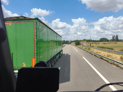 The image shows a perspective from inside a vehicle, looking out at a highway scene. A large green truck with a yellow tarp on top is traveling on the right side of the road. The truck has multiple axles and a red stripe along its side. A yellow road sign is visible in the distance to the right, indicating directions to Montpellier and Valence, and also mentioning "Suivre Paray LL M." The sky is blue with fluffy white clouds. The road is paved, with white lane markings. The surrounding environment appears rural, with fields and trees. The weather is sunny. The cropped images provide closer views of the truck's details, the road sign, and the surrounding landscape.
