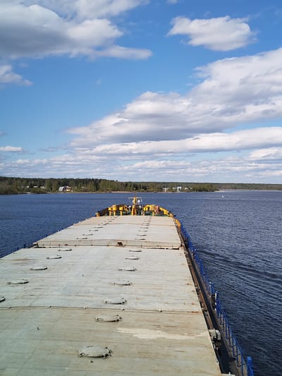 A long, flat-topped barge is centered, cutting through a body of water, with a view from the stern towards the bow. The barge is gray with a series of oval hatches along its surface and a blue railing along its right side. At the bow, there are yellow and black structures. The water is dark blue with gentle ripples. Across the water is a line of trees and some visible structures, suggesting a shoreline. The sky is blue with scattered white clouds. The location is Irdomatka, Russia.