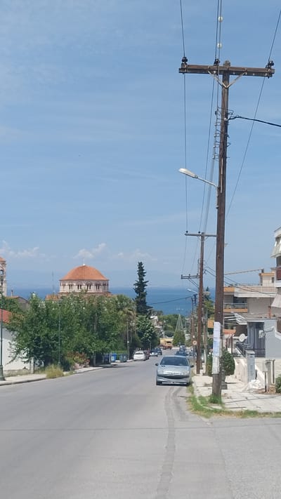 The image depicts a street scene in Nea Michaniona, Greece, under a clear blue sky with scattered white clouds. The perspective is from a slightly elevated position, looking down a paved road that slopes towards a body of water, likely the sea.
On the right side of the road, a prominent wooden utility pole stands tall, adorned with numerous wires and insulators, characteristic of urban infrastructure. A street light is attached to this pole. Adjacent to the pole, there are buildings with a modern architectural style, featuring balconies and white facades.
On the left side of the road, a distinct building with a terracotta-colored dome, topped with a cross, is visible in the mid-ground. This architectural feature suggests a church or a similar religious structure. Lush green trees and foliage surround this building, creating a contrast with the urban elements.
A silver hatchback car is parked on the right side of the road, facing away from the camera. Further down the road, several other vehicles are present, including what appears to be a yellow truck or van and a few other cars, indicating moderate traffic.
The background reveals a distant shoreline with hills or mountains and the expanse of the sea. The overall scene conveys a typical Mediterranean urban environment on a sunny day.