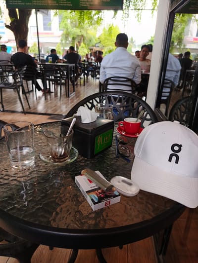 A medium shot captures a table at an outdoor cafe in Pantegajah, Indonesia. The table, made of a dark, speckled glass material, is cluttered with several items. To the left, a clear glass of water sits next to a small, empty glass on a saucer with a spoon inside, which appears to have contained a dark liquid, likely coffee. In the center, a black tissue box and a red teacup with a saucer containing a frothy, light brown beverage are visible. Resting on the table are a pack of cigarettes, a lighter, a pair of glasses, and a white baseball cap with a black logo. In the background, patrons are seated at tables, some facing away from the camera, and lush green foliage is visible, suggesting an outdoor or semi-outdoor setting. The overall atmosphere appears relaxed, with people engaged in casual conversation and enjoying their drinks.
