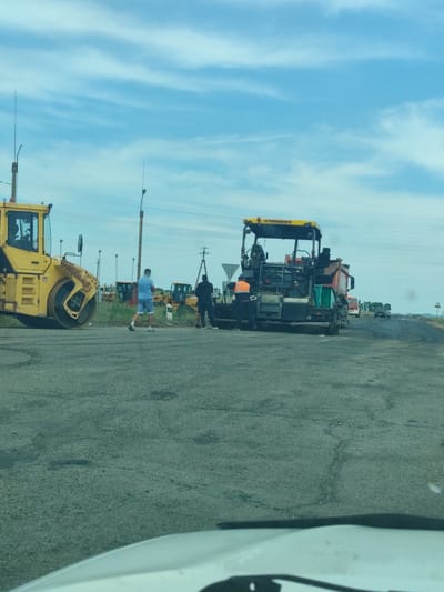 The image depicts a road construction scene in Beya, Russia. The foreground shows a grey asphalt road surface, possibly under repair or construction, with some cracks visible. In the middle ground, a yellow Bomag roller is parked on the left, and a VOGELE paving machine with an attached truck is actively laying asphalt on the right. Two men are visible near the paving machine, one in a blue shirt and shorts walking away from the camera, and another in an orange vest and dark pants standing on the paving machine. In the background, more construction vehicles, including excavators, are parked. Utility poles with power lines stretch across the scene, and a few trees are sparsely scattered in the distance. A dark car is driving on the road further down. The sky is partly cloudy, suggesting daytime with potentially fair weather. The overall impression is of an ongoing infrastructure project. The visible text on the paving machine reads "VOGELE".