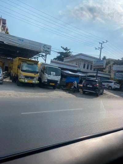 The image depicts a roadside view in Mandar Sari, Indonesia. In the foreground, a paved road with a white line marking runs across the bottom. In the mid-ground, several vehicles are parked. Two yellow and white dump trucks are prominent, with the yellow truck facing the viewer, displaying license plate "KH 8339 LH". Next to it is a white Isuzu ELF truck, also facing the viewer, with a "DIJUAL" (for sale) sign on its windshield. Further to the right, a black SUV with license plate "DA 1549 HO" is parked, and behind it, a white SUV with a red and white circular logo.
A building with a blue and white striped awning is visible behind the trucks. A sign above the building reads "MIRAKULUM MOTOR" and lists services such as "BODY REPAIR SERVICE SPARE PART". Power lines crisscross the sky, and a utility pole is visible on the right side of the frame. Trees are scattered in the background. The sky is bright blue with some scattered clouds, indicating daytime. The overall scene suggests a commercial area or a vehicle dealership.