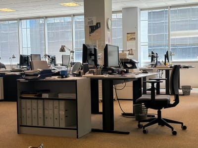 This image depicts a bright, open-plan office environment in San Sebastián De Los Reyes, Spain, during daytime. The scene is dominated by multiple workstations, each featuring computer monitors, keyboards, office chairs, and desk lamps. Large windows along the back wall are fitted with horizontal blinds, allowing diffused natural light to illuminate the space. The ceiling consists of a grid of square tiles with integrated fluorescent lighting.
In the foreground, a desk includes a side shelf unit containing numerous white binders, some with visible labels, along with stacks of documents and office supplies. A black office chair with wheels is positioned next to this desk. Other desks further back display similar setups with multiple monitors, printers, and organized papers.
A central pillar within the office features several notices. One prominent sign reads "EMERGENCIA 0307851", and another poster displays the text "TODO VUELVE" (Everything Returns). A wall calendar is visible on another pillar, showing "PANADERIA LÓPEZ" and the month "NOVIEMBRE" (November), indicating a Spanish-speaking context.
The office appears largely unoccupied, with only one person partially visible on the far left, facing away from the camera. Despite the absence of many people, the workstations are fully equipped and appear ready for use, suggesting a quiet moment during the workday or after hours. The space is functional and tidy, reflecting a professional working environment.
