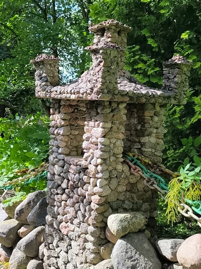 The image shows a miniature stone castle structure, likely a garden ornament or folly, set against a backdrop of lush green foliage. The castle is intricately constructed from various sizes and colors of natural stones, with multiple turrets and a roof. A thick, multi-colored chain, predominantly green and white, is wrapped around the base of the structure, partially obscured by the surrounding rocks and plants. The ground is covered with more rocks, some of which are quite large and irregular in shape. The overall impression is of a whimsical, handmade creation nestled within a natural outdoor setting. The provided context indicates this scene is located in the city of Mosėdis, Lithuania, known for its stone museum and natural stone formations.
