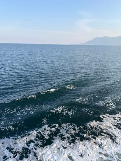 The image presents a wide-angle marine vista near Kato Karioutes, Greece, captured under a clear, pale blue sky with a subtle atmospheric haze. The scene is dominated by dark blue-green sea water, extending from the immediate foreground to a distant horizon. The water surface in the mid-ground features small waves and ripples, with scattered points of sunlight reflection. The lower right quarter of the frame exhibits a distinct area of white foam and turbulent water, indicative of a vessel's wake. A distant, hazy, light grey mountainous landmass is visible on the right side of the horizon line. No individuals or other specific man-made objects are depicted within the frame.