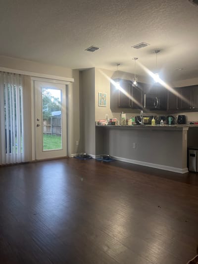 The image depicts the interior of a residential living area adjacent to a kitchen, with a view of a backyard through a glass door. The flooring is dark hardwood. In the foreground, the floor shows some smudges and reflections from overhead lighting. To the left, a glass door leads to a backyard with a wooden fence and green grass, partially obscured by vertical blinds. In the middle ground, a portion of the kitchen counter is visible, with dark cabinets above it. The counter is stocked with various items including bottles, containers, and kitchen appliances like a coffee maker and a black air fryer. There are three pendant lights hanging from the ceiling above the kitchen counter. On the floor near the kitchen counter, two pet bowls are placed on blue mats. To the right of the kitchen counter area, a silver trash can is visible. The walls are painted a neutral color. The image does not contain any information that would allow for determination of its specific location within Jacksonville, United States of America.