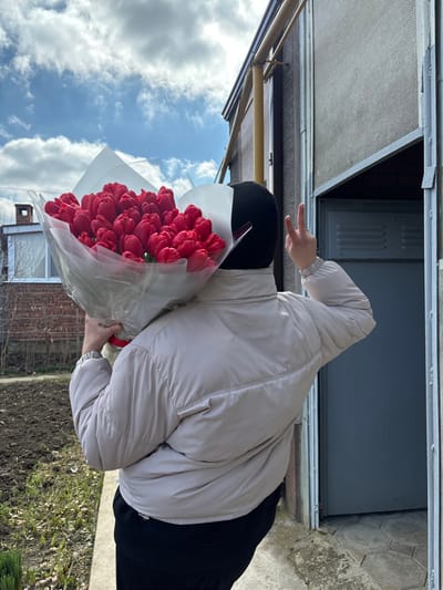 An outdoor, eye-level photograph depicts an individual from behind, standing on a concrete pathway. The individual wears a black balaclava, a light beige puffer jacket, and dark-colored pants. They hold a large bouquet of numerous red tulips, wrapped in clear plastic film and white paper, with their left arm. Their right hand is raised, forming a V-sign with the index and middle fingers. To the right, a light brown building with dark upper siding features a partially open doorway or window with a visible metal frame and an obscured, dark interior. In the left background, a red brick building with a white-framed window and a chimney is visible. The sky is blue with scattered white clouds. The ground immediately adjacent to the concrete pathway is bare earth with sparse, nascent green plant growth. This scene is located in Trudobelikovskiy, Russia.