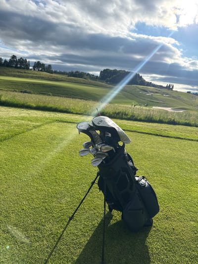 An outdoor, eye-level, vertical, medium shot captures a golf bag filled with clubs standing on a manicured golf course. The golf bag, primarily black, is positioned on a vibrant green tee box. Several golf clubs, with silver heads and white covers, are visible protruding from the bag. The background features rolling green hills, a well-maintained golf course with sand traps, and a line of trees along the horizon. The sky above is a mix of dramatic, dark clouds and bright, sunlit patches, with a sunbeam cutting diagonally across the frame. The scene is set during daylight, with the sun casting shadows on the grass. The location is identified as a golf course in Kløfta, Norway, implied by the characteristic landscape and golfing activity. No people are prominently featured in the immediate foreground; however, several figures are faintly visible on the distant golf course. The overall atmosphere is one of a serene, yet potentially dynamic, day for golf.
