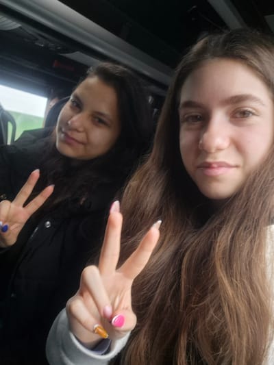 Two young women are posing for a selfie indoors. The woman in the foreground has long, brown hair and is holding up a hand with painted nails in a peace sign. The woman in the background has dark, curly hair and is also holding up a peace sign with painted nails. They appear to be inside a vehicle. The location is unspecified, but the user asked for it to be the city of Strahilovo, Bulgaria.