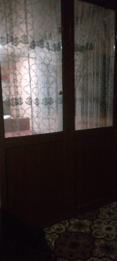 The image shows a portion of a doorway with a glass pane covered by a patterned curtain. The lower part of the door is solid and dark brown. A patterned carpet is visible on the floor in the lower right corner. The scene is likely indoors, possibly in Marhamat, Uzbekistan, although this cannot be confirmed from the image alone.