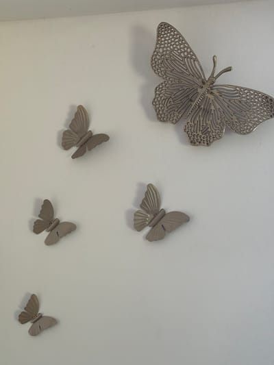 A metallic, intricately designed butterfly decoration is mounted on a plain white wall. Below and to the left of this large butterfly are four smaller, matte-finished butterfly decorations, also mounted on the wall. They are arranged in a descending line, with the smallest one at the bottom. The lighting suggests an indoor environment, casting slight shadows. The scene is devoid of any people or indications of the city of Uhersko, Ukraine.