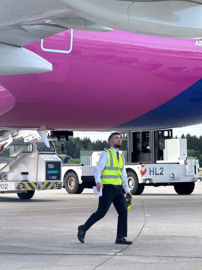 A man wearing a white shirt, dark tie, and a high-visibility yellow vest is walking on an asphalt tarmac in front of a large airplane. The airplane has a distinctive pink and blue livery. The man is carrying a yellow item in his right hand and appears to be looking up at the airplane. Behind him, a white vehicle, possibly a tow tractor for the aircraft, is visible. The vehicle has markings including "FP02", "TLD ELECTRIC", and "power stow". Another white vehicle with the marking "HL2" is also present. The scene is outdoors, with trees and a cloudy sky in the background. The location is identified as Künersberg, Germany.