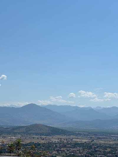 The image captures a wide aerial view of the town of Gornji Kokoti, Montenegro, under a clear blue sky dotted with a few fluffy white clouds. The foreground is dominated by the lush green foliage of trees and bushes, obscuring the immediate lower portion of the frame. Beyond this, the landscape unfolds into a tapestry of rolling hills and a sprawling residential area characterized by red-tiled roofs scattered amongst greenery. Further in the distance, a distinct, rounded hill stands prominently, its slopes covered in sparse vegetation. The background is a majestic range of layered mountains, their peaks fading into a hazy blue silhouette against the horizon, suggesting a vast and mountainous terrain surrounding the town. The scene conveys a sense of peaceful rural life nestled within a dramatic natural landscape.