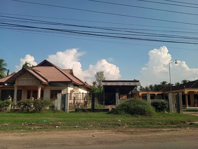 The image depicts a street scene in Lhoknga, Indonesia, on a clear day with a blue sky dotted with white, fluffy clouds. In the foreground, a dirt road with some scattered debris runs horizontally across the bottom of the frame. Beyond the road is a strip of green grass, also with some litter.
The middle ground features several structures. On the left is a beige house with a reddish-brown tiled roof, featuring a covered porch supported by pillars. A black metal fence encloses the property, with lush green bushes and flowering plants visible behind it. To the right of this house is an open-sided structure with a corrugated metal roof, likely a carport or workshop, where a dark-colored vehicle is parked. Another building, possibly a mosque or community hall, is visible further to the right, characterized by its long, covered arched walkways and a similar reddish-brown tiled roof. A street lamp stands tall in front of one of the buildings. Palm trees are scattered throughout the background, suggesting a tropical or subtropical environment. Several power lines stretch across the top of the image, intersecting the sky. No people are clearly visible or identifiable in the scene. The overall impression is one of a residential or community area in a coastal town in Indonesia.