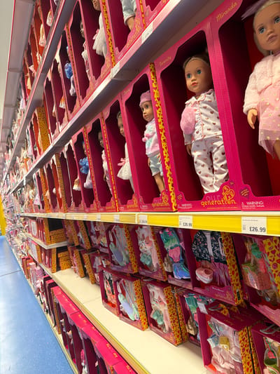 The image shows shelves stocked with "Our Generation" dolls and related accessories, suggesting a retail setting, likely a toy store or department store. Price tags of £26.99 are visible. The dolls are packaged in pink boxes with clear windows displaying the doll and its attire. The names "Miranda" and "Serenity" are visible on the doll packaging. The doll outfits and accessories are packed in yellow boxes. This is possibly located in a city, based on the presence of retail goods. Considering the currency (GBP), it's likely in the United Kingdom, and there is no way to determine it is in Wrexham with certainty.