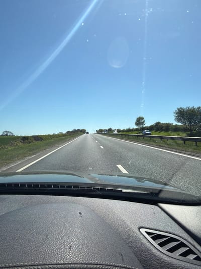 The image, originally rotated 90 degrees clockwise, shows a view from inside a vehicle looking forward through the windshield onto a road. The scene depicts a bright, clear day, likely late morning or afternoon, given the bright blue sky and visible sun glare.
The road, a wide asphalt surface with distinct white lane markings, stretches into the distance. A broken white line is visible in the middle, suggesting multiple lanes of traffic or a separated carriageway. To the left of the road is a green grassy verge, separated from the road by a metal guardrail. Beyond the guardrail, trees and bushes line the landscape, indicating a rural or semi-rural setting. Several vehicles, including what appears to be a white van and other cars, can be seen in the distance on the road.
The upper left portion of the image is dominated by a bright, clear blue sky, showing significant lens flare and small bright specks, possibly dust on the windshield or lens artifacts. The right side of the image features the interior of the car, including a dark grey or black textured dashboard or door panel with an air vent visible in the upper right. The edge of the windshield and its frame are also discernible.
The overall impression is of a drive on an open highway or main road on a sunny day in the area of Northallerton, United Kingdom. No discernible text is visible in the image.