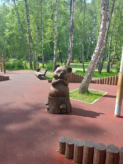 The image depicts a children's playground in a park setting. The foreground features a dark red rubberized surface, typical of modern playgrounds, with several wooden log obstacles arranged in a semi-circular fashion. In the center of the playground, a whimsical wooden sculpture of a pig sitting on a tree stump is prominently displayed. The pig sculpture has detailed features, including large ears and a snout, and appears to be carved from a single piece of wood. Behind the sculpture, a wooden park bench is visible, situated on a patch of green grass.
The background is dominated by a dense forest of birch trees, characterized by their distinctive white bark with black markings. The trees are in full leaf, suggesting a summer or late spring setting, and sunlight filters through the canopy, casting dappled shadows on the ground. Some elements of an adventure park or rope course, such as ropes and wooden structures, can be seen in the further background, indicating that this playground is part of a larger recreational area. A white pole, possibly a part of the playground equipment or a park amenity, stands on the right side of the image. The overall atmosphere is one of a natural, recreational space designed for children's activities.
This playground is located at the "Park of Legends" in Chelyabinsk, Russia.