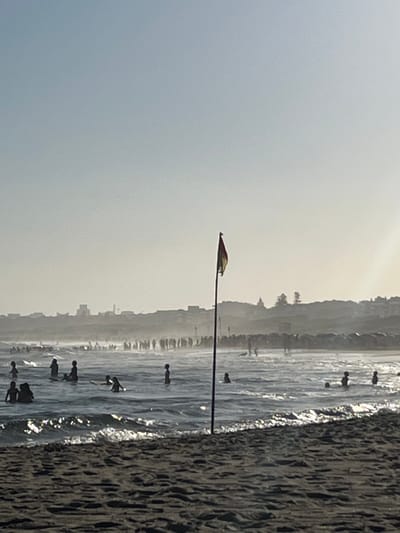 A sideways view of a beach featuring a calm ocean with surfers in the water. The coastline is visible in the background, with buildings and trees. A lamp post stands near the shore. The sand is textured with the impression of waves. The sky is hazy.
