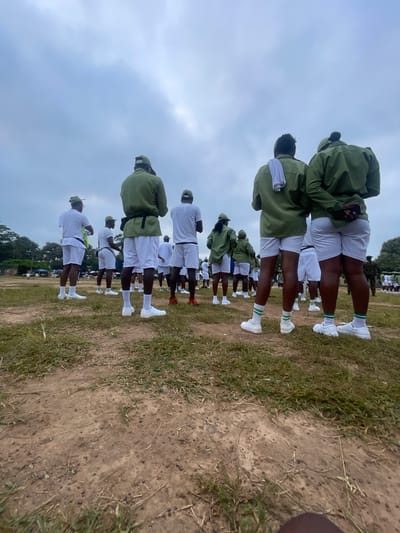 This media file captures a group of young adults, likely participants in a national service program given their uniform, standing in formation outdoors. The subjects are predominantly seen from behind, dressed in matching olive green jackets and white shorts, paired with white socks and athletic shoes. They are arranged in several rows, with the perspective shifting throughout the image, suggesting a rotating camera or a series of shots.

The scene appears to be a parade ground or open field with patches of grass and dirt. In the background, hints of trees and possibly some temporary structures suggest a rural or semi-urban training environment. The sky is overcast, indicating a cloudy day. The overall mood seems to be one of order and discipline, with the subjects standing attentively.

The image shows several individuals, with the focus shifting from the front of the group to the back and then to the sky. The lighting suggests it's daytime, likely morning or afternoon, due to the even light and lack of harsh shadows. There is no visible text in the image. The context of Emure-Ekiti, Nigeria, points to a potential orientation or training camp for youth service members in that region.