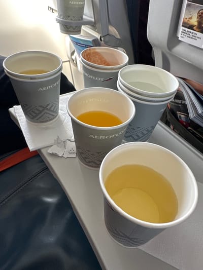 The image depicts a tray table on an airplane, likely in economy class, with four cups of beverages. Three of the cups are partially filled with liquid, one with a bright orange juice, and two with lighter yellow liquids, possibly tea or water. The fourth cup is empty and stacked on top of another empty cup, suggesting these are for use. The cups are grey with a geometric pattern and the "Aeroflot" logo. There is also a white paper napkin and a small packet of sugar. In the background, a magazine is visible with a picture of a man's face and Russian text. The seats in front and beside are visible, one of which is upholstered in dark material. The scene is well-lit, likely by interior cabin lighting, and the overall impression is one of a standard in-flight service. There is no specific indication of the location being the city of Sadovoe, Russia, but given Aeroflot is the national airline of Russia, it is plausible.