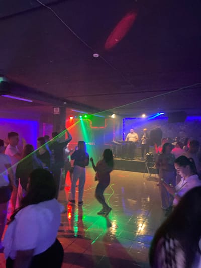The image depicts a dimly lit nightclub in Nisporeni, Moldova, with a crowd of people dancing. Laser lights, in green and red hues, cut through the smoky atmosphere, illuminating the dance floor. In the background, a DJ booth is visible with a person tending to the equipment, likely a keyboard. The floor is reflective, catching the colored lights. The overall scene suggests a lively and energetic nightlife atmosphere.
