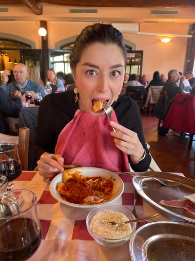 The image depicts the interior of a restaurant in Civitavecchia, Italy. The setting is characterized by a rustic aesthetic, featuring wooden ceiling beams, light-colored walls, and warm illumination from wall-mounted fixtures. Tables are uniformly covered with red and white checkered tablecloths.

In the foreground, a woman with dark hair pulled back is centered, mid-action, bringing a piece of light-colored food to her widely open mouth with a fork held in her right hand. She wears a black long-sleeved top and a large pink napkin tucked into her collar. A gold-colored bracelet is visible on her right wrist, and a distinctive light-colored earring is present on her left ear. Her left hand holds a knife near her plate.

On the table in front of her, a white bowl contains partially consumed pasta with a red meat sauce. To her immediate right, a small glass bowl holds grated Parmesan cheese with a spoon. Further right, a silver platter presents slices of cured ham, with another empty silver platter beneath it. Two glasses filled with red wine are positioned to the left.

The background reveals a bustling dining area with numerous other patrons seated at various tables. An older man with a bald head, wearing a light blue jacket, is visible to the left. Other individuals of varying ages are seen dining and conversing. Some chairs in the background feature red fabric drapes. Air conditioning vents are mounted on the upper walls.