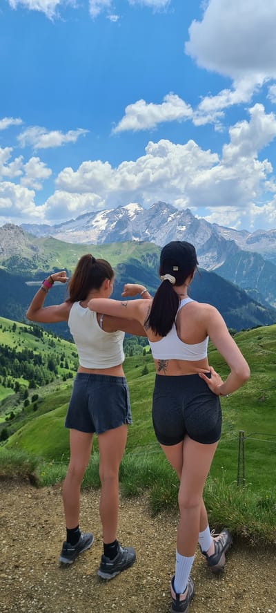 Two women stand on a mountain path, their backs to the camera, overlooking a vast mountain range under a blue sky with scattered clouds. The woman on the left wears a white tank top and dark grey shorts, flexing her biceps. The woman on the right wears a white sports bra and dark grey shorts, with her left hand on her hip. Both women are wearing athletic shoes. The background features lush green hills dotted with trees, leading up to snow-capped mountains in the distance. The location is the city of Sëlva - Wolkenstein - Selva di Val Gardena, Italy.