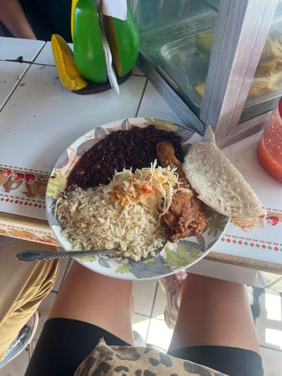 The image shows a close-up, overhead view of a plate of food, likely at an outdoor eatery or food stall in Managua, Nicaragua. The plate is filled with a meal consisting of white rice, black beans, a piece of fried chicken, and a small portion of coleslaw. A folded tortilla rests on top of the chicken. Next to the plate, to the left, are what appear to be sliced green plantains and a banana, suggesting a side dish or dessert. In the background, a glass display case with more food items is visible, along with a small bowl of red sauce and a patterned placemat. The presence of sunlight and the casual setting suggest it is daytime. The composition hints at a person eating, with parts of their legs and torso visible in the foreground, one wearing black shorts and a leopard-print top, and the other wearing tan pants and blue sneakers. No visible text is present in the image.