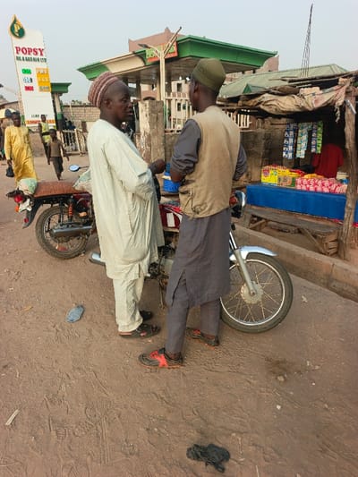 Two men in traditional clothing stand next to a red motorcycle, conversing on a dusty road. A gas station sign reading "POPSY OIL & GAS NIG LTD" is visible in the background. A small roadside stall selling snacks and beverages is situated to the right, under a makeshift awning. A man in a yellow robe walks by in the distance, with another child walking in the same direction. The ground is uneven, showing signs of wear.