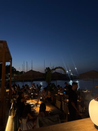 An outdoor waterfront restaurant is depicted at dusk or early night within a marina in Heraklion, Greece. The sky is dark blue, transitioning to a lighter hue near the horizon. Numerous patrons, predominantly adults, are seated at wooden tables with place settings, engaged in dining and conversation. Each table is illuminated by a small, glowing portable lamp. Large, circular thatched parasols are positioned above some dining areas. Several restaurant staff members are present, with one male in a dark uniform visible prominently on the right. In the background, across a body of water, multiple sailboats with visible masts are moored in the marina. A large, brightly illuminated white motor yacht is also discernible further out, casting reflections on the water. Wooden structural elements of the restaurant's canopy are visible on the left, and a large, spherical white lamp is partially visible in the bottom right foreground.