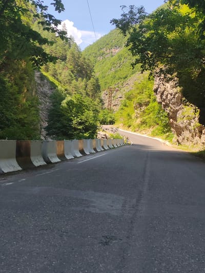 The image depicts a scenic, winding road through a mountainous gorge in the region of Ajara, Georgia. The road is paved with asphalt and features a concrete barrier along its left side, which shows signs of age and weathering. The gorge walls are steep and covered with lush green vegetation, including trees and shrubs. Rocky outcrops are visible on the right side of the gorge. The sky is blue with a few white clouds. In the distance, a transmission tower can be seen. The overall impression is one of natural beauty and a remote, mountainous landscape.