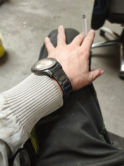 A close-up shot focuses on a person's right arm, adorned with a metallic wristwatch and clad in a grey, ribbed-cuff jacket. The person is holding a lit cigarette between their fingers, with a wisp of smoke rising upwards. Dark pants cover the lower portion of the arm, and a portion of a black bag with a zipper is visible. The background consists of a concrete floor and a metallic chair. The scene appears to be outdoors, potentially in Volevčice, Czechia, given the concrete ground and industrial-style chair.