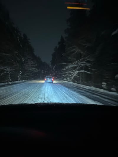 A nighttime view from a vehicle traveling on a snow-covered road. Sparse trees line the roadside, dusted with snow. Headlights illuminate the road ahead, revealing tire tracks in the snow. In the distance, the taillights of another vehicle are visible. The scene is dimly lit, with the dominant colors being white, gray, and black, creating a stark contrast. The focus is on the road and the car in front, highlighting winter driving conditions.