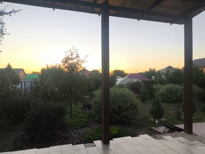 The image depicts a backyard scene at dusk, viewed from under a wooden overhang supported by a dark brown pillar. The sky transitions from a pale blue at the top to a warm yellow-orange near the horizon. In the middle ground, various trees and shrubs are visible, some with a dark foliage and others lighter green. A small tree with orange berries is positioned in the left-center of the frame. Behind the vegetation, several houses with pitched roofs are discernible, along with a greenhouse. The foreground features light-colored paving stones, some of which appear to be laid out, suggesting ongoing construction or landscaping. The scene suggests a residential area in a rural or suburban setting, likely in Мичурин ауылдық округі, Kazakhstan, given the context. There are no people or animals present in the image.