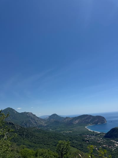 The image depicts a landscape in Grabovica, Montenegro. The photograph is oriented vertically. The sky, a gradient of deep blue, dominates the left side of the image. The right side of the image contains a series of mountains covered in green vegetation. Between the mountains and the sky is a body of water, which appears to be the Adriatic Sea. A coastal town is nestled along the edge of the water, with the presence of buildings suggested by clusters of structures partially obscured by vegetation. The overall environment suggests a mountainous, coastal region under clear, sunny conditions.