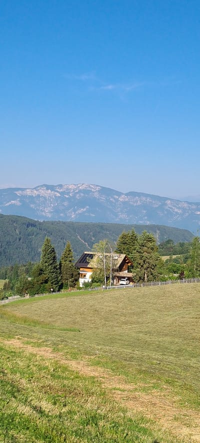 The image depicts a sunlit, rural landscape under a clear blue sky. In the foreground, a mown grassy field slopes gently upwards towards a cluster of trees and a house. The field exhibits lines from recent mowing, with patches of cut grass drying in the sun.

Dominating the middle ground is a house with a pitched roof, adorned with solar panels on one section. The house is nestled amongst mature evergreen trees and some deciduous trees. A white car is parked under a sheltered area adjacent to the house. A wooden fence runs along the property line.

In the background, a dense forest covers the lower slopes of mountains. The mountains themselves form a distant, hazy backdrop, their peaks and ridges detailed and rocky, suggesting a rugged alpine environment.

The scene appears tranquil and idyllic, characteristic of a countryside setting. The location is identified as Deutschnofen - Nova Ponente, Italy, suggesting the image was captured in this specific mountainous region. There are no people visible in the image, and no discernible actions are taking place. The focus is on the natural scenery and the architectural elements within it.