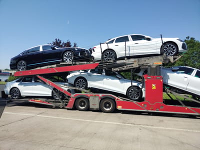 The image shows a car carrier truck loaded with several sedans. On the top level, a dark colored sedan is parked on the left, and a white sedan is parked on the right. Below them, on the lower level, two white sedans are visible, with one parked on the left and another on the right, partially obscured by the carrier structure. The truck is red and appears to be parked on a concrete surface, possibly a parking lot or road. The background consists of a clear blue sky and some green trees. There are no people visible in the image. The overall scene depicts a transport of vehicles. Based on available information, this scene is likely located in Oberbeisheim, Germany.