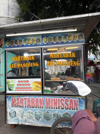 This media file depicts a street-side food stall in Lampeuneurutblang, Indonesia. The stall appears to be selling various types of martabak and kue pancong, as indicated by the prominent text on its signage. The stall itself is a mobile cart, likely a common sight in bustling street markets.

The primary subjects are the food stall and its offerings. The stall features glass windows displaying food items, including what looks like martabak and kue pancong, as well as rows of canned goods, possibly condensed milk or other ingredients. The signage, predominantly in yellow and red, is in Indonesian. Visible text includes "MARTABAK," "KUE PANCOENG," and "MARTABAK MINISSAN." The side of the stall has a blue and white banner with hexagonal images, possibly depicting ingredients or finished products.

In the background, glimpses of the surrounding environment can be seen. There are other street vendors, a few motorbikes, and people moving around, suggesting a busy street scene. A person's arm and shoulder, wearing a purple top, are visible in the foreground, indicating the viewer is likely on a motorbike or standing very close to the stall, perhaps as a customer. A motorcycle mirror is also visible, reflecting the sky and surroundings.

The lighting suggests it might be daytime, possibly overcast given the muted light. The overall impression is of a vibrant, everyday street scene in Indonesia, focused on local food commerce. There are no strong emotional cues, but the image conveys a sense of lively commerce and daily life.