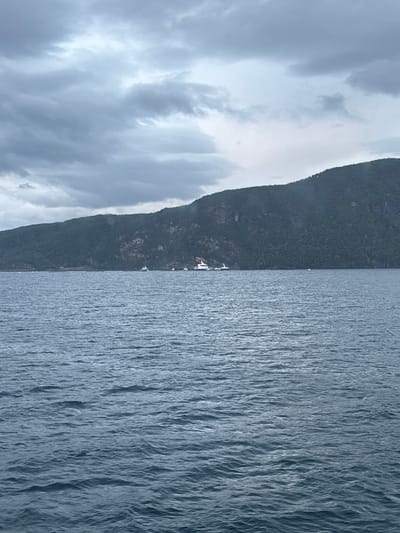 A wide shot captures a body of water with a mountainous, tree-covered shoreline in the background. The sky is overcast with heavy clouds, suggesting a cool or potentially rainy day. Several boats are visible on the water, including a large white ferry or passenger boat, and smaller vessels. The water is choppy, with visible waves. The scene appears to be a natural landscape, possibly a fjord, with human elements of transport and recreation. The location is identified as Russevåg, Norway. There are no people visible in the image.