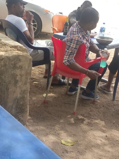 Several people sit around a plastic table outdoors. One man wears a white shirt and baseball cap and another has on a plaid shirt. A bottle of Guinness and drinking water sits on the table, along with a metal bowl. The people sit in plastic chairs, red and blue. The ground is dirt, and a single leaf lies on the ground. A car is visible in the background.