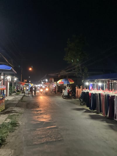 The image depicts a street scene at night in Proyonangg, Indonesia, likely a marketplace. The street is paved and appears wet, reflecting the ambient light from overhead lamps and shop stalls. On the left side of the street, there are several stalls with awnings, displaying goods and illuminated by artificial lights. Some people are visible walking along the sidewalk and in front of the stalls.

In the middle of the street, there are several motorcycles, some parked and others in motion, their headlights creating streaks of light. People are also seen walking or standing in the street. Colorful umbrellas, possibly used to shield from sun or rain, are set up at some stalls.

On the right side of the street, more stalls are visible, one of which displays a rack of clothing, likely pants, hanging on hangers. This stall is covered by a blue awning and illuminated by lights, making the merchandise visible. More motorcycles are parked near this stall. The overall impression is of a bustling night market or a street lined with shops open late. The sky is dark, indicating it is nighttime, and some overhead power lines are visible. A lone tree with foliage is also present in the background.