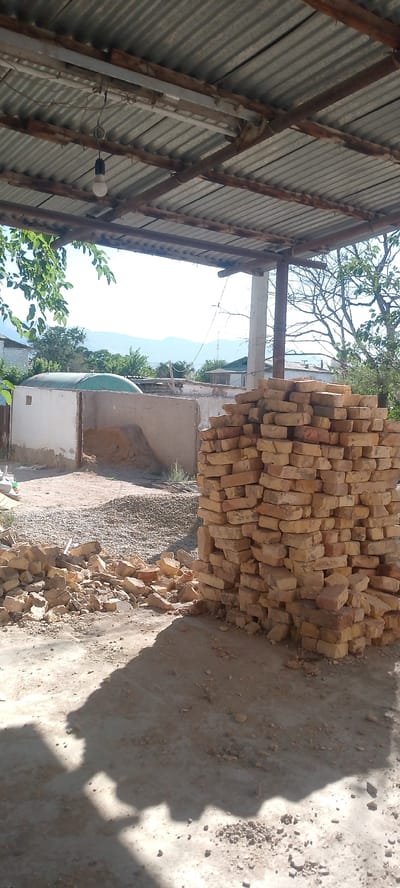 The image depicts an outdoor scene in Balkanabat, Turkmenistan, likely on a construction site or a storage area. A corrugated metal roof supported by wooden beams and a concrete pillar is visible overhead. A bare light bulb hangs from the ceiling. In the foreground, a large pile of yellow bricks stands prominently, with scattered bricks and debris on the ground around it. Behind the bricks, there's a low, weathered concrete wall. Beyond the wall, a large, greenish, dome-shaped structure is partially visible, possibly a water tank or silo. Further in the background, residential buildings and trees are scattered across the landscape, with mountains faintly visible on the horizon under a bright, clear sky. The ground is dusty and appears to be a mixture of dirt and gravel. The scene is bathed in sunlight, casting distinct shadows on the ground. No people are visible in the image.