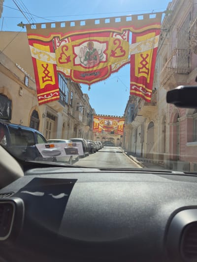 The image captures a street scene in the city of Birżebbuġa, Malta, as viewed from inside a vehicle. Large, ornate banners are strung across the street, featuring red and yellow fabric with intricate designs and religious imagery. The banners appear to be celebrating a local event or festivity, possibly religious, given the depiction of a saint on one of them. The architecture of the surrounding buildings is characteristic of Maltese towns, with traditional stone facades and balconies. The street is lined with parked cars, and the sky is clear and blue, indicating a sunny day. The foreground shows a portion of the vehicle's interior, including the dashboard and windshield, with reflections from the outside visible. The overall impression is one of a vibrant cultural celebration taking place in a historic Maltese setting.