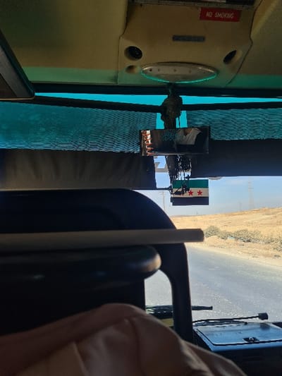 A daytime photograph captures the interior of a vehicle looking out onto an arid landscape along a paved road, identified as being in Al-Kiswa, Syria.

**Vehicle Interior:** The upper portion of the image shows the vehicle's beige ceiling, featuring an oval light fixture, two circular vents, and a red "NO SMOKING" sign with white text. Below the ceiling, a dark teal pleated curtain or shade partially covers the top of the windshield. A rearview mirror hangs centrally, reflecting a person wearing a white shirt. The lower frame reveals part of a black dashboard or seat structure, with a light-colored cylindrical object (resembling a cigarette or pen) resting horizontally on it. In the bottom right, a person's forearm, clad in light pink/peach fabric, is partially visible.

**Exterior Environment:** Through the windshield, an asphalt road extends into the distance under a clear, bright sky. The surrounding terrain is dry and arid, characterized by sandy-brown hills and sparse, low-lying green vegetation. In the middle distance, utility poles and power lines are visible against the horizon. A Syrian flag, with black, white, and green horizontal stripes and two red stars on the white stripe, is clearly visible, seemingly mounted on an external structure near the road, possibly a signpost or checkpoint.