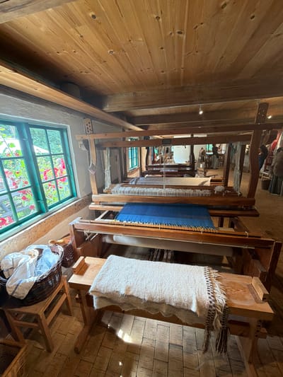 In the city of Lejre, Denmark, this image captures the interior of a room with a large, wooden loom as the central subject. The loom is actively being used, with blue fabric partially woven. To the left of the loom, a green-framed window looks out onto a garden with red flowers. A wicker basket filled with white fabric sits on a wooden stool next to the window. A white woven blanket with a patterned fringe is draped over a wooden bench in front of the loom. The room has a rustic ambiance with exposed wooden beams and a wooden floor. In the background, other looms and weaving equipment are visible, suggesting a workshop or a museum exhibit dedicated to textile arts. Several individuals are present in the background, appearing to be engaged in activities related to the looms or observing the process. The lighting is natural from the window and artificial from ceiling lights, creating a warm and inviting atmosphere.
