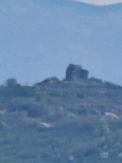 The image shows a rocky hilltop in the city of Kumanovo, North Macedonia. Atop the hill is a distinct, darker rock formation that could be a part of a building or a natural structure. The foreground of the image displays a slope covered with shrubs and vegetation, suggesting a natural environment. The background reveals a blurry sky, further implying a distant perspective.