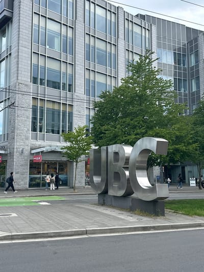 The image depicts a modern, multi-story building with a glass and grey facade, likely located in an urban environment. In front of the building stands a large, brushed metal sculpture of the letters "UBC", partially obscured by a tree. Several people are walking on the sidewalk in front of the building, and some are gathered near a storefront labeled "SHO PPER'S" (presumably "Shoppers Drug Mart" with partially covered text). A portion of a street is visible next to the sidewalk. The general aesthetic suggests a university campus setting. Considering that Electoral Area A, Canada, encompasses the University of British Columbia, it is highly probable that this scene is located on the UBC campus. No interactions are visible.