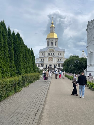 The image depicts a view of the Holy Trinity Cathedral in Diveyevo, Russia, from a paved walkway. The cathedral, with its prominent golden dome and cross, is situated at the end of the path. A dense line of manicured evergreen trees runs along the left side of the walkway, separating it from a lower hedge. Several people are walking along the path towards the cathedral. Some are dressed in traditional modest clothing, with women wearing headscarves. In the foreground, a couple walks away from the camera. The sky is overcast with dramatic clouds. The right side of the image shows a white building with arched windows, likely part of the monastery complex. Flowers are visible in a garden bed along the right edge of the walkway.